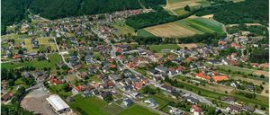 Luftaufnahme einer kleinen Stadt mit vielen Häusern, grünen Feldern und einer Straße. Berge und Bäume umgeben die Stadt.