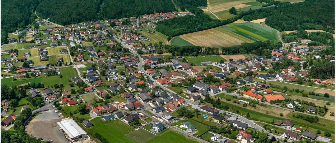 Luftaufnahme einer kleinen Stadt mit vielen Häusern, grünen Feldern und einer Straße. Berge und Bäume umgeben die Stadt.