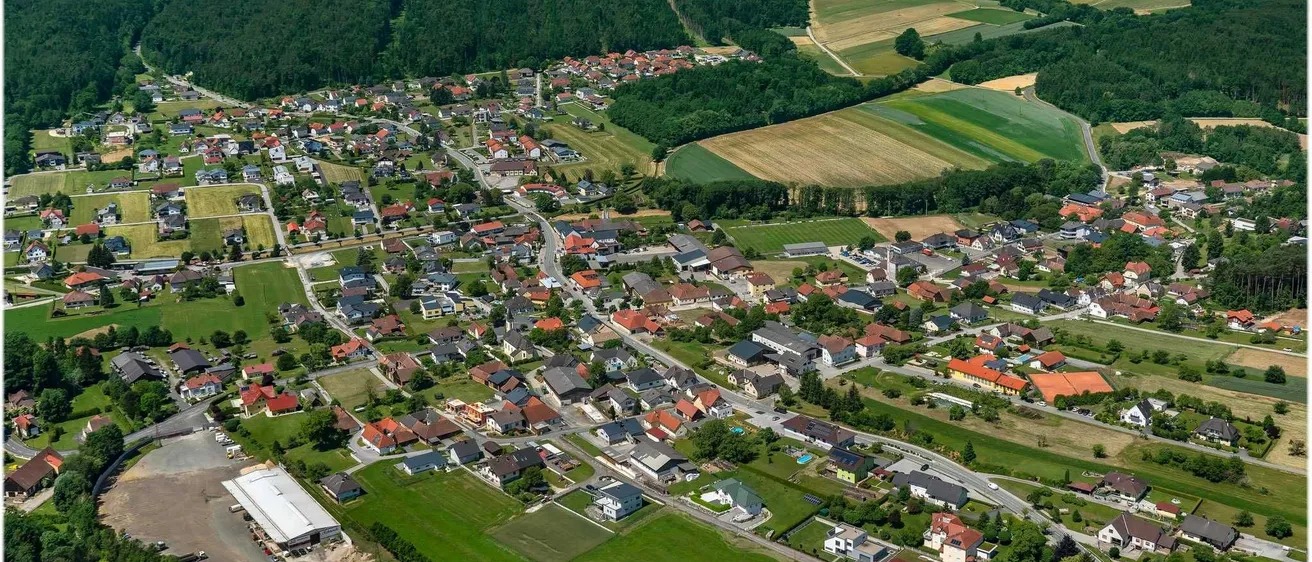 Luftaufnahme einer kleinen Stadt mit vielen Häusern, grünen Feldern und einer Straße. Berge und Bäume umgeben die Stadt.