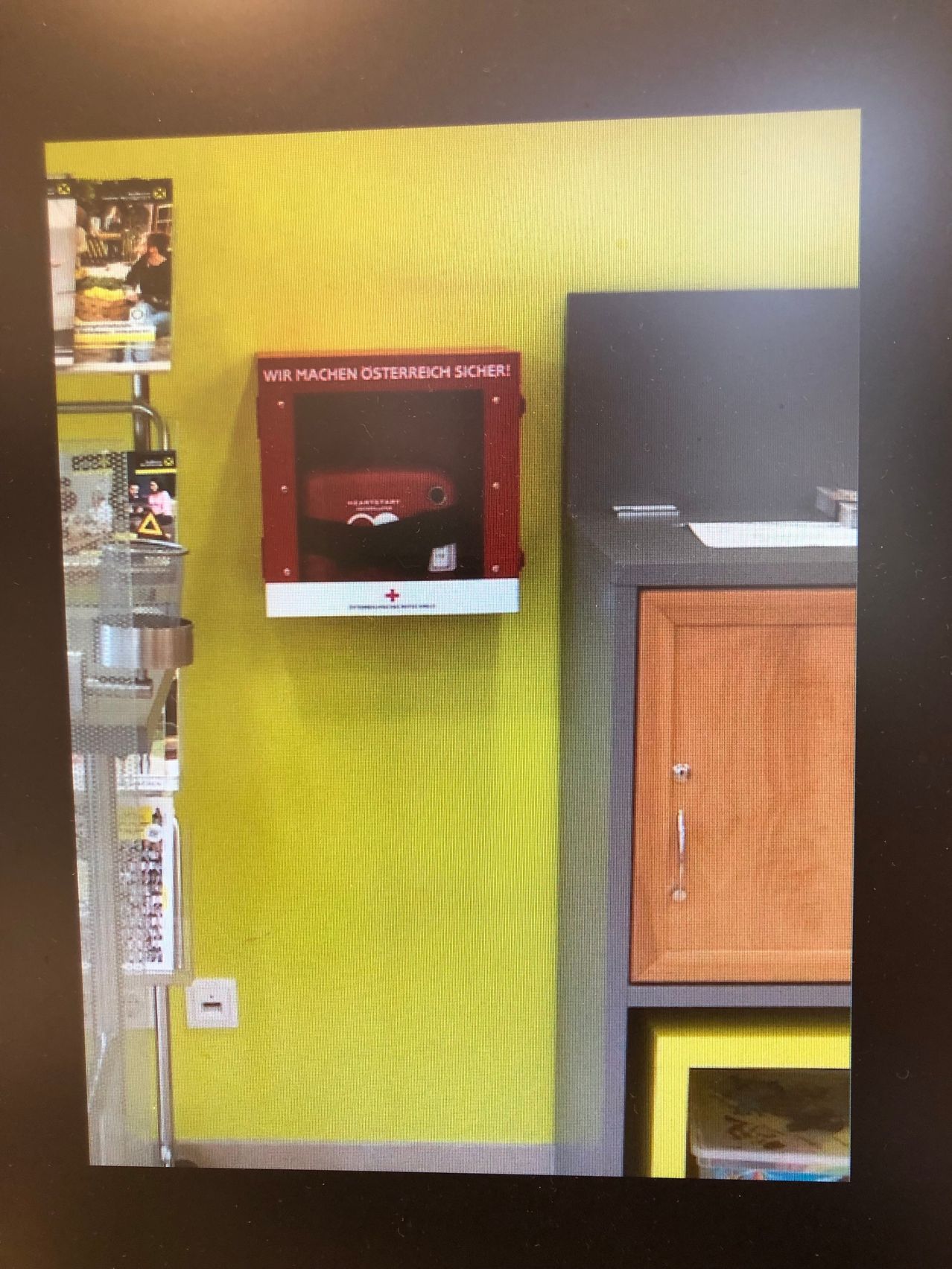 Eine gelbe Wand mit einer montierten roten Box, die 'WIR MACHEN OSTERREICH SICHER!' besagt, mit einem medizinischen Gerät im Inneren. Ein grauer Regal und ein hölzerner Schrank mit einem silbernen Griff sind ebenfalls sichtbar.