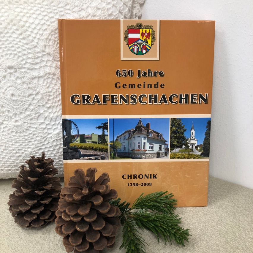 Ein Buch mit dem Titel '650 Jahre Gemeinde Grafenschachen Chronik 1358-2008' steht auf einem Tisch mit Tannenzapfen und Zweigen. Der Einband zeigt ein Wappen und Bilder eines Hauses, einer Kirche und eines Ortes.
