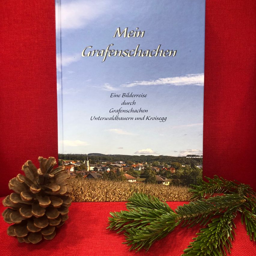 Ein Buch mit dem Titel 'Mein Grafenschachen' wird auf einem roten Tuch präsentiert, mit einem Tannenzapfen und Tannenzweigen in der Nähe. Das Cover zeigt eine ländliche Landschaft mit Gebäuden unter einem blauen Himmel.