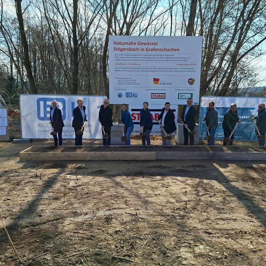 Eine Gruppe von Menschen mit Schaufeln posiert für ein Foto vor einem Schild, das 'Naturnahe Gewässerspeicherung in Grafenschachen' mit einem Budget von 1.552.000 Euro anzeigt.
