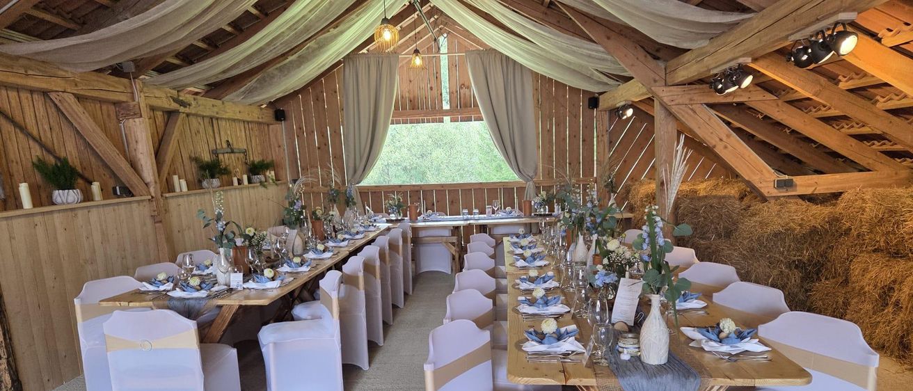 Ein rustikaler Scheunenraum für eine Veranstaltung mit langen Tischen, weißen Stühlen und dekorativen Blumen und Kerzen unter Holzbalken und Vorhängen.
