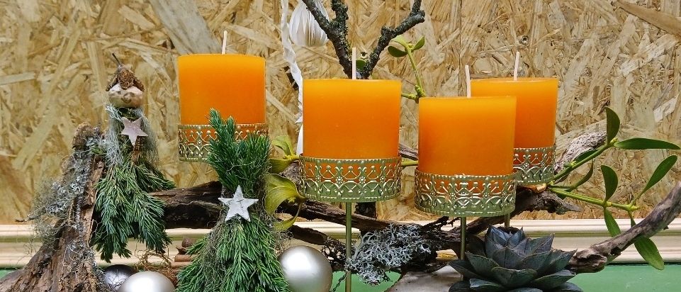 Eine Holzplattform zeigt vier orangefarbene Kerzen mit grünen Haltern, begleitet von einem Weihnachtsbaum, silbernen Ornamenten und einer Sukkulente. Der Hintergrund zeigt eine Holzwand.