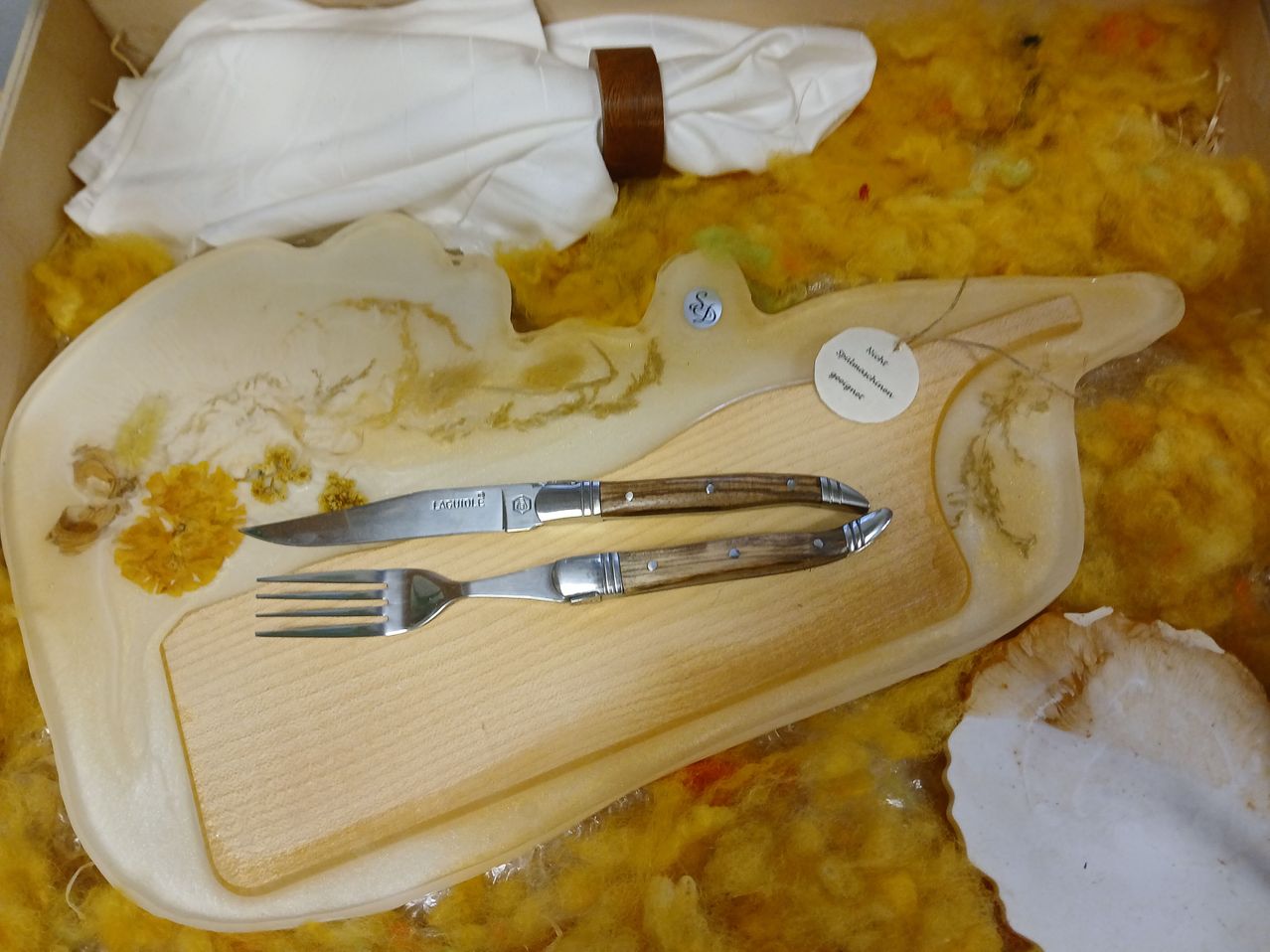 Ein Holzschneidbrett zeigt ein Messer und eine Gabel mit Holzgriffen. Ein gefalteter weißer Servietten ist obenauf, und ein kleines Schild mit Text ist angebracht. Der Hintergrund zeigt ein gelbliches Material mit einigen weißen und gelben Objekten.