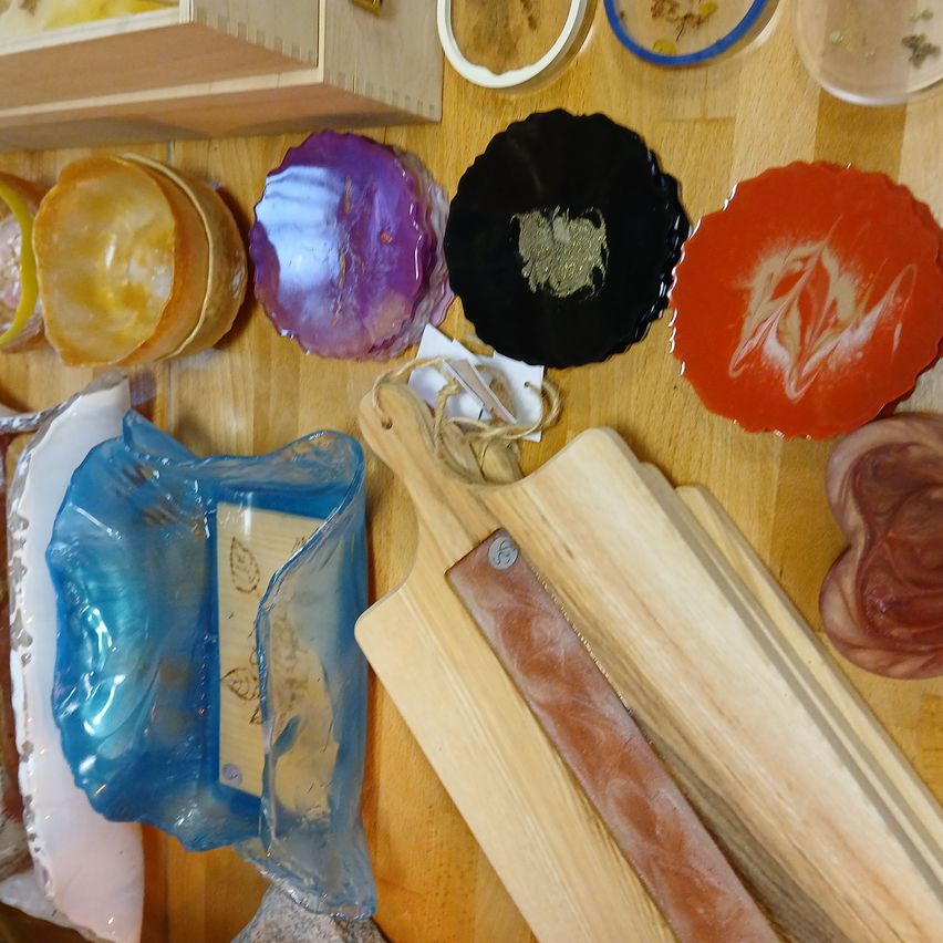 Ein Holztisch zeigt verschiedene Kunstwerke, darunter farbige Teller, Schüsseln und eine blaue Glasschale. Zwei Holzschneidebretter mit Ledergriff sind ebenfalls zu sehen.