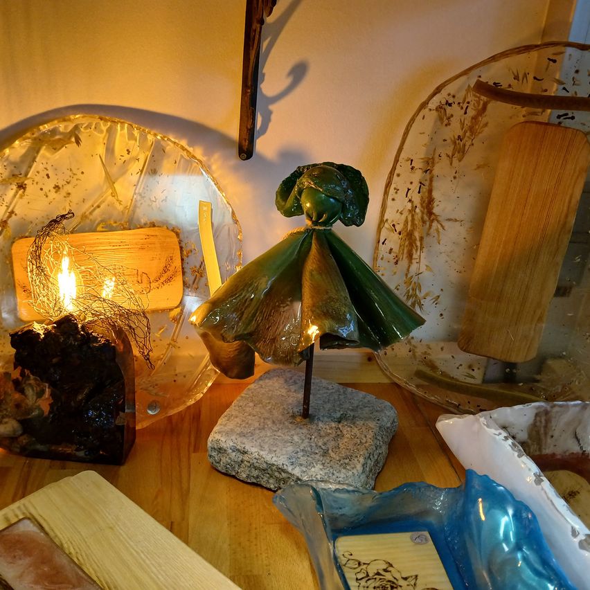 Ein Holztisch zeigt verschiedene Dekorationsartikel. Eine kleine, grüne, blattartige Figur steht auf einem grauen Stein. In der Nähe sind eine Glasschale mit einer brennenden Kerze und eine Glasplatte mit einem Holzstab zu sehen. Andere Glasschalen mit verschiedenen Designs sind ebenfalls vorhanden.