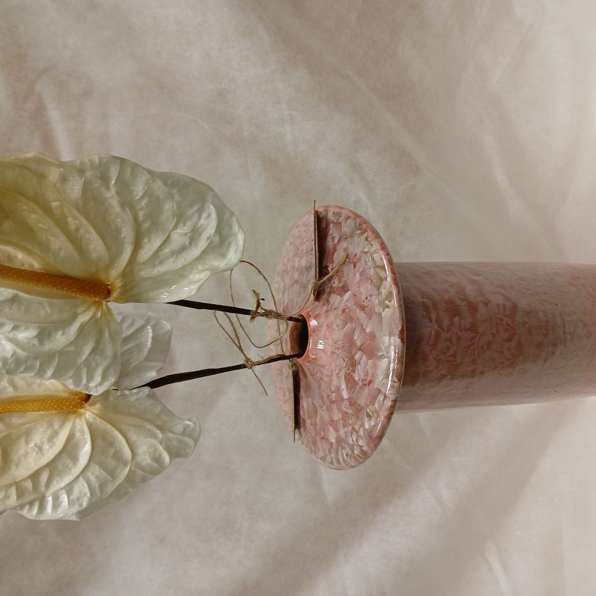 Ein Nahaufnahme einer weißen Blume mit langen Stielen in einer rosa Keramikvase mit strukturierter Oberfläche, auf einem weißen Tuchhintergrund platziert.