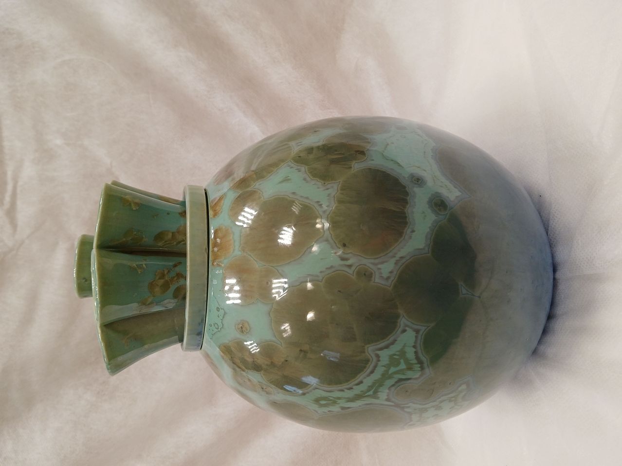 Eine runde Keramikvase mit grünem und braunem Blumenmuster auf weißem Hintergrund. Die Vase hat zwei grüne Griffe an den Seiten.