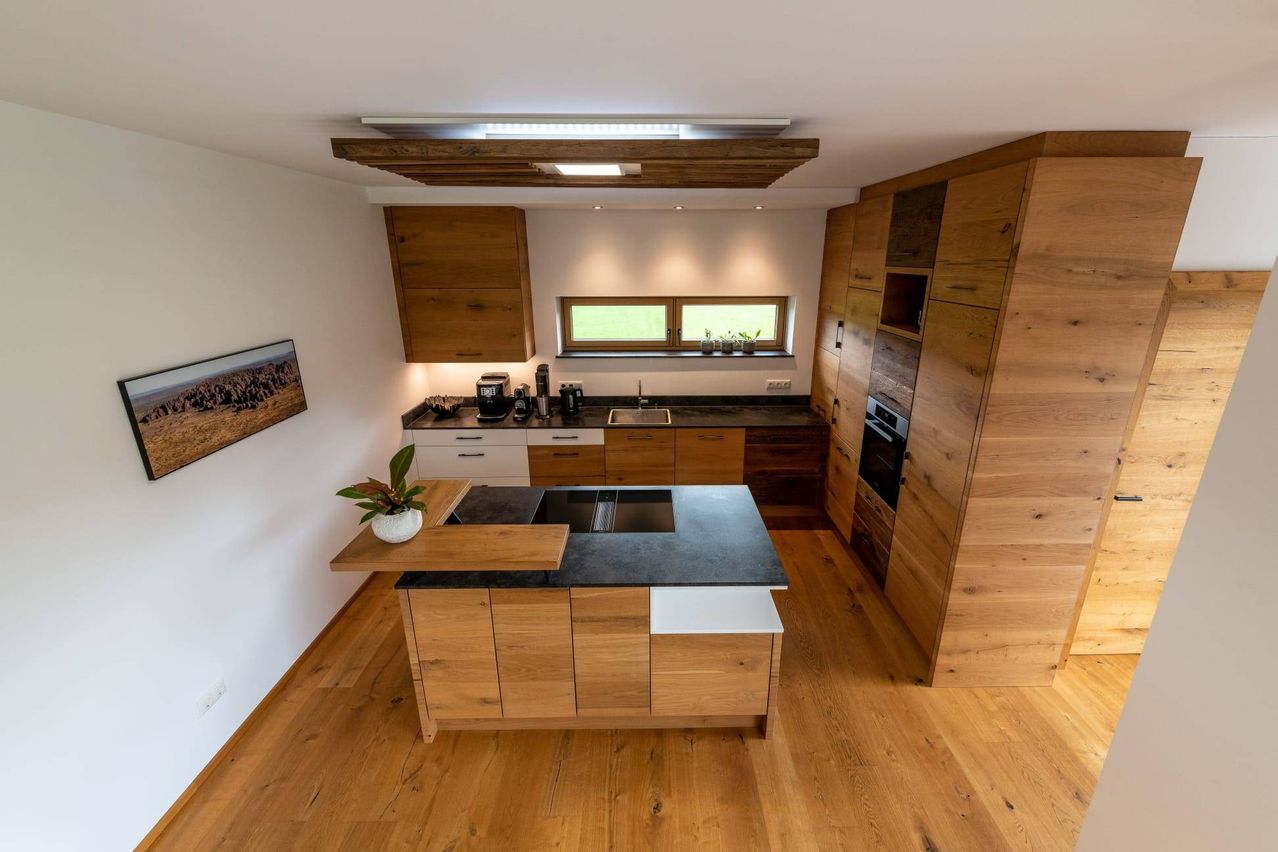 A modern kitchen with wooden cabinets, a black countertop, and a large window with plants on the sill.