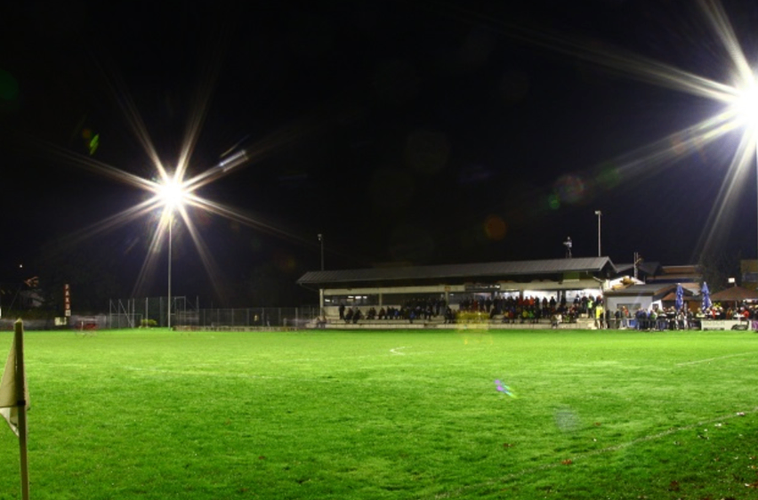 Ein Fußballfeld mit hellen Lichtern, die die Nacht erhellen, mit Leuten auf den Tribünen, die zuschauen.