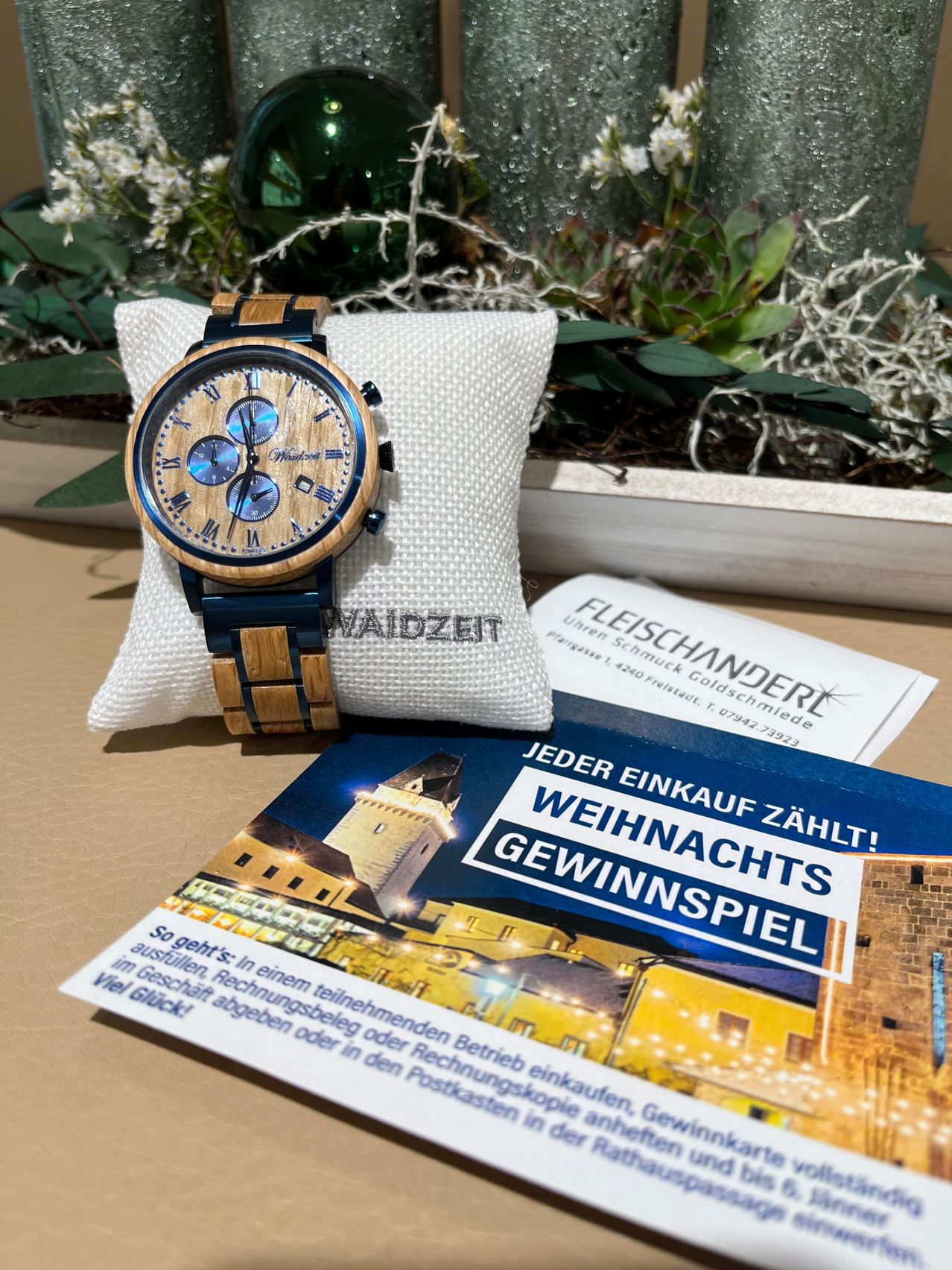 Eine hölzerne und blaue Armbanduhr auf einem weißen Kissen mit einem Prospekt, der 'Weihnachtszählung! Jeder Kauf zählt! Weihnachtsraffle' und eine Burg im Hintergrund zeigt.