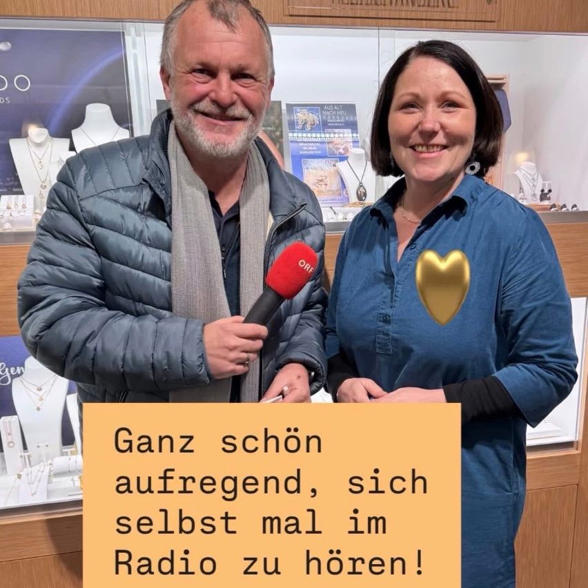 Ein Mann und eine Frau stehen in einem Juwelierladen, lächelnd. Der Mann hält ein Mikrofon und trägt einen Schal. Die Frau hat ein goldenes Herz auf ihrem Shirt. Sie stehen vor einer Vitrine mit Schmuck. Ein gelbes Schild mit deutschem Text steht vor ihnen.