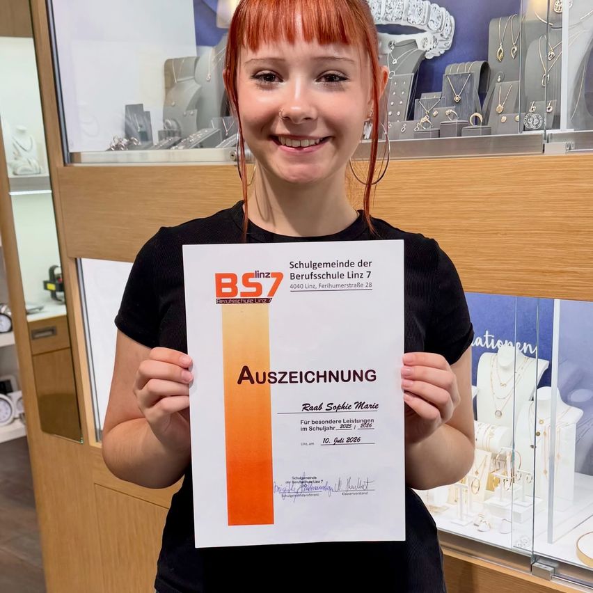 Eine Frau mit roten Haaren hält ein Zertifikat vor einer Schmuckvitrine. Das Zertifikat lautet 'Auszeichnung' und stammt von der Berufsschule Linz 7.
