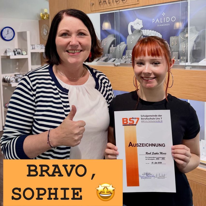 Zwei Frauen stehen in einem Juwelierladen, eine hält ein Zertifikat mit dem Text 'BRAVO, SOPHIE' und einem Stern-Emoji. Die Frau auf der linken Seite gibt einen Daumen hoch. Dahinter ist eine Vitrine mit Schmuck.