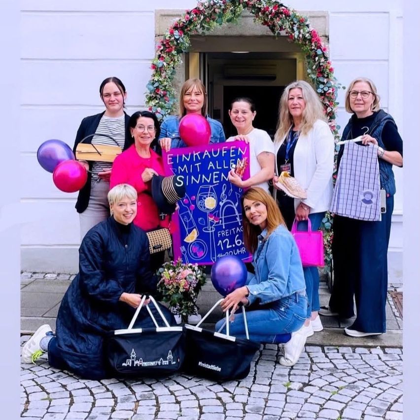 Eine Gruppe von Frauen steht vor einem Gebäude mit Luftballons. Sie halten ein Banner, Taschen und Blumen. Eine hält ein Holzbrett, eine andere einen rosa Luftballon.