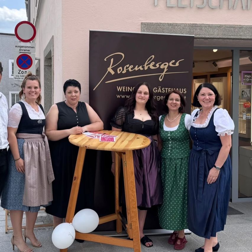 Fünf Frauen stehen vor einem Geschäft mit einem Schild, das 'Rosenkegler' steht. Sie sind in traditioneller Kleidung gekleidet und lächeln für ein Foto.