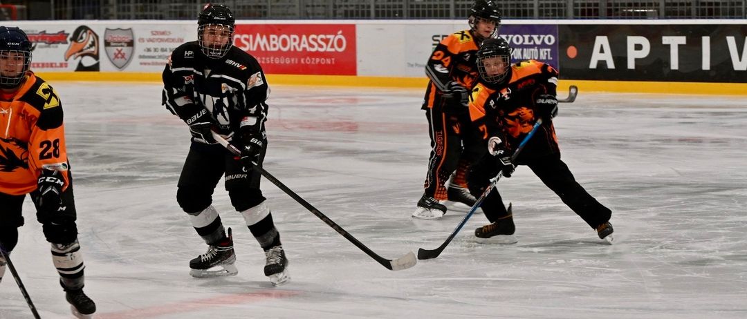 Drei Eishockeyspieler gleiten auf dem Eis und halten Eishockeyschläger. Sie tragen schwarze, weiße und orangefarbene Uniformen. Ein Spieler trägt einen schwarzen Helm. Hinter ihnen zeigt eine Wand Werbung, darunter ein roter Banner mit weißem Text.