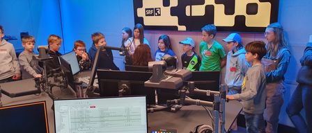 Eine Gruppe von Kindern steht hinter einem Schreibtisch in einem Radiostudio und beobachtet einen Monitor mit Text und Zahlen. Sie scheinen etwas über Radiosendungen zu lernen.