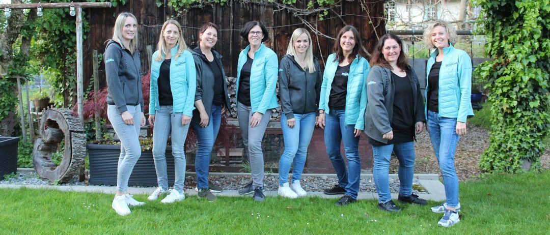 A group of women wearing blue jackets and jeans are posing for a photo in front of a wooden house.