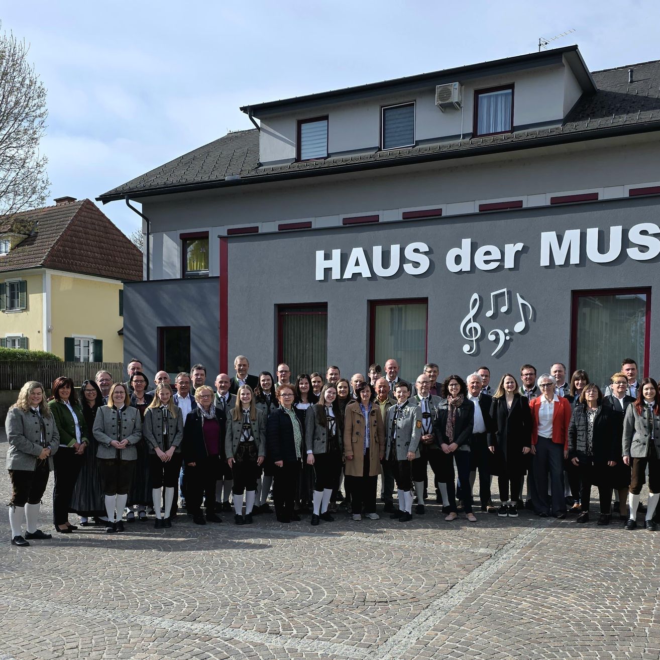 Eine Gruppe von Menschen in traditioneller deutscher Kleidung posiert vor einem Gebäude mit 'HAUS der MUS' auf der Fassade.