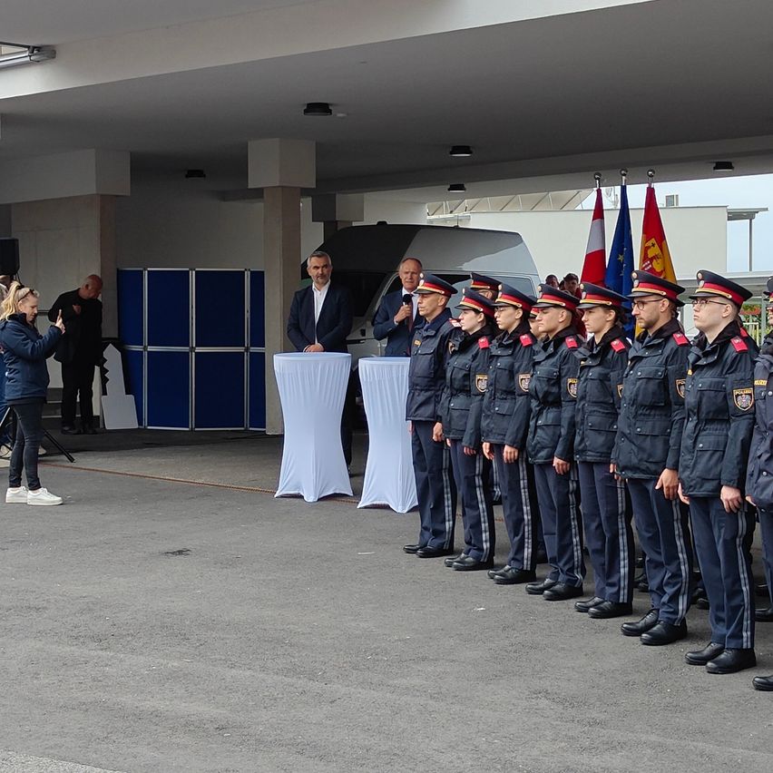 Eine Gruppe von Polizeibeamten steht in einer Halle in einer Reihe, während ein Mann an einem Podium spricht. Flaggen sind hinter ihnen angebracht.