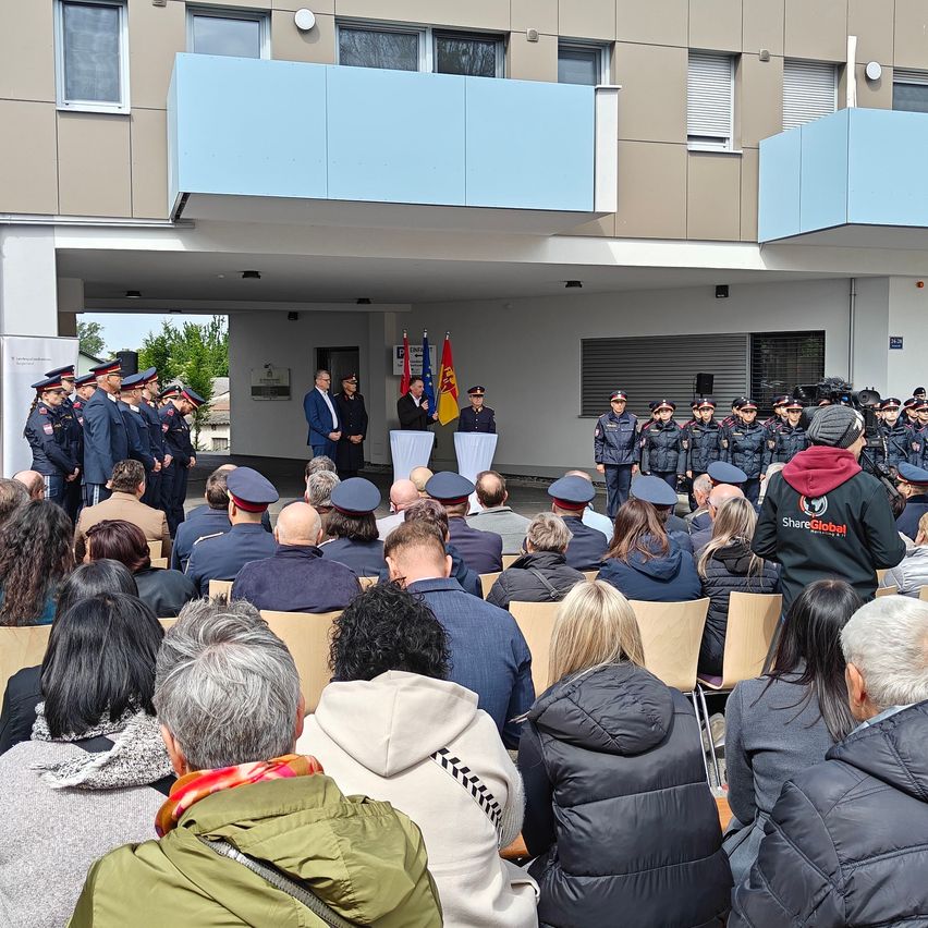 Eine Veranstaltung im Freien zeigt uniformierte Personen, einschließlich Polizeibeamter, vor einem Gebäude mit Fahnen. Viele Teilnehmer, einige mit Mützen, sitzen auf Stühlen und beobachten den Ablauf.