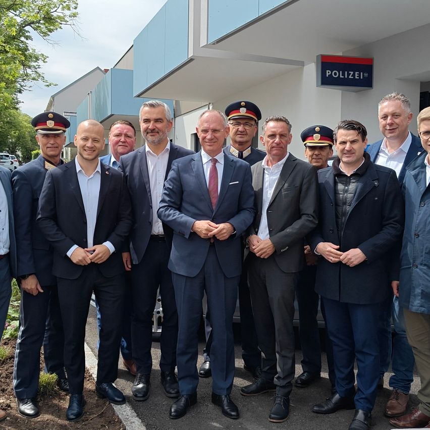 Eine Gruppe von Männern in Anzügen, Hüten und Krawatten steht vor einem Polizeigebäude. Bäume und ein Schild mit der Aufschrift Polizei sind im Hintergrund zu sehen.