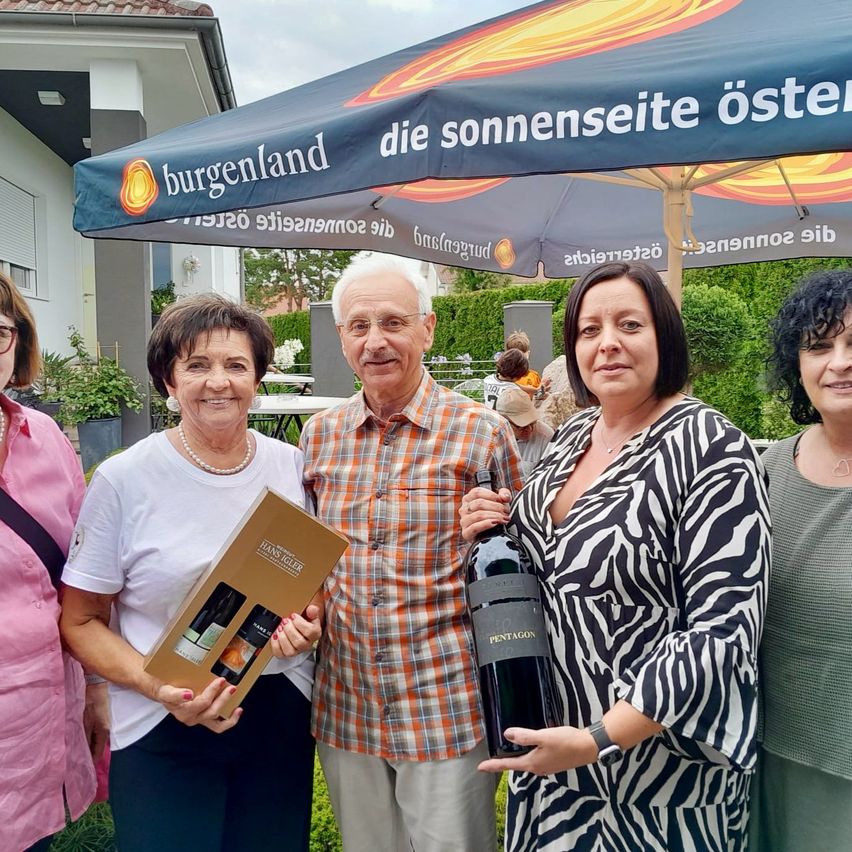Eine Gruppe von vier Personen steht unter einem Regenschirm und lächelt für ein Foto. Eine Frau hält eine Weinflasche und eine Weinbox, während eine andere Frau eine Flasche hält. Dahinter steht ein Gebäude mit einem Haus und Bäumen.
