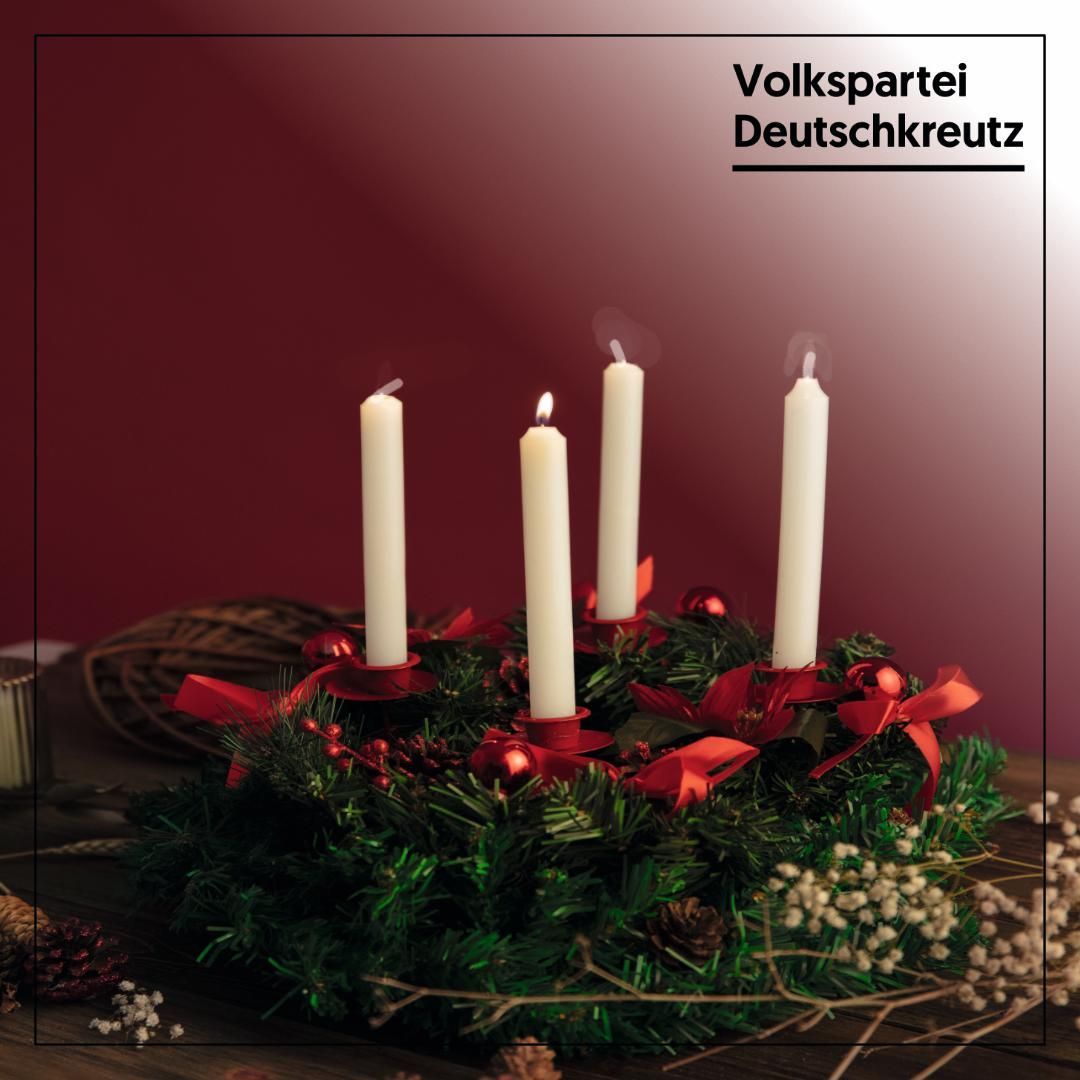 Ein Weihnachtskranz mit vier brennenden Kerzen, geschmückt mit roten Bändern und Tannenzapfen, auf einem Holztisch vor einem kastanienbraunen Hintergrund.