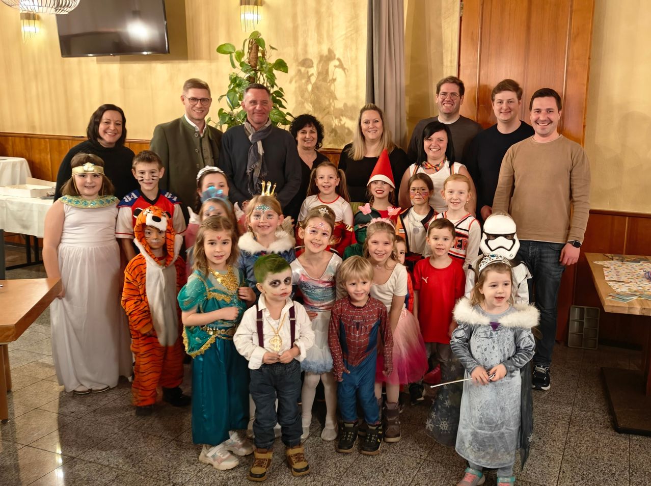 A group of adults and children dressed in various costumes pose for a photo in a room. The adults include both men and women, and the children are wearing a range of outfits, including a tiger costume, a spider-man costume, and a Star Wars Stormtrooper costume.