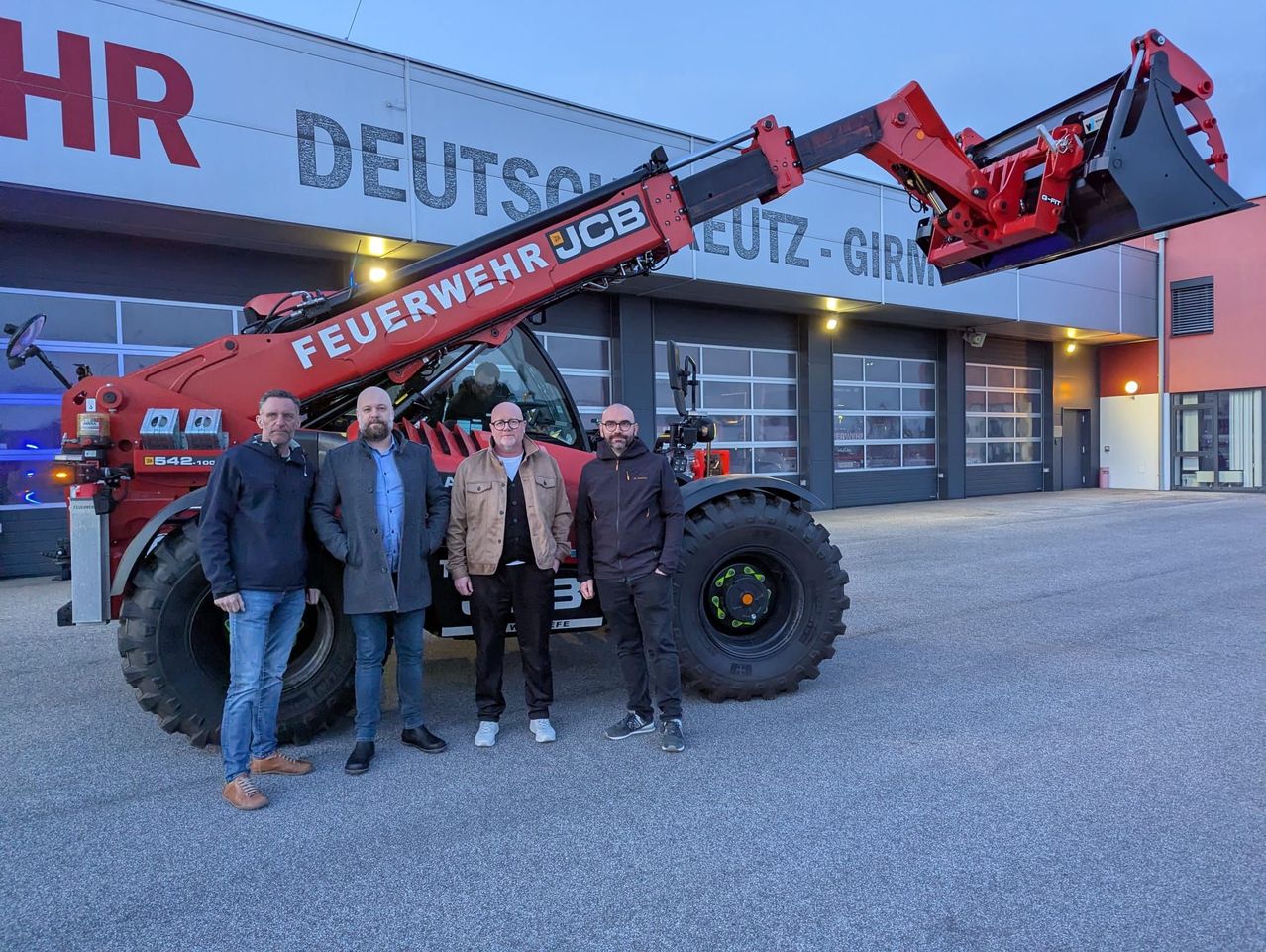 Four men stand in front of a red JCB machinery vehicle, posing for a photo. The vehicle has the words 'FEUERWEHR JCB' written on it. Behind them is a building with large windows and a sign that reads 'DEUTSCH-GIR'.
