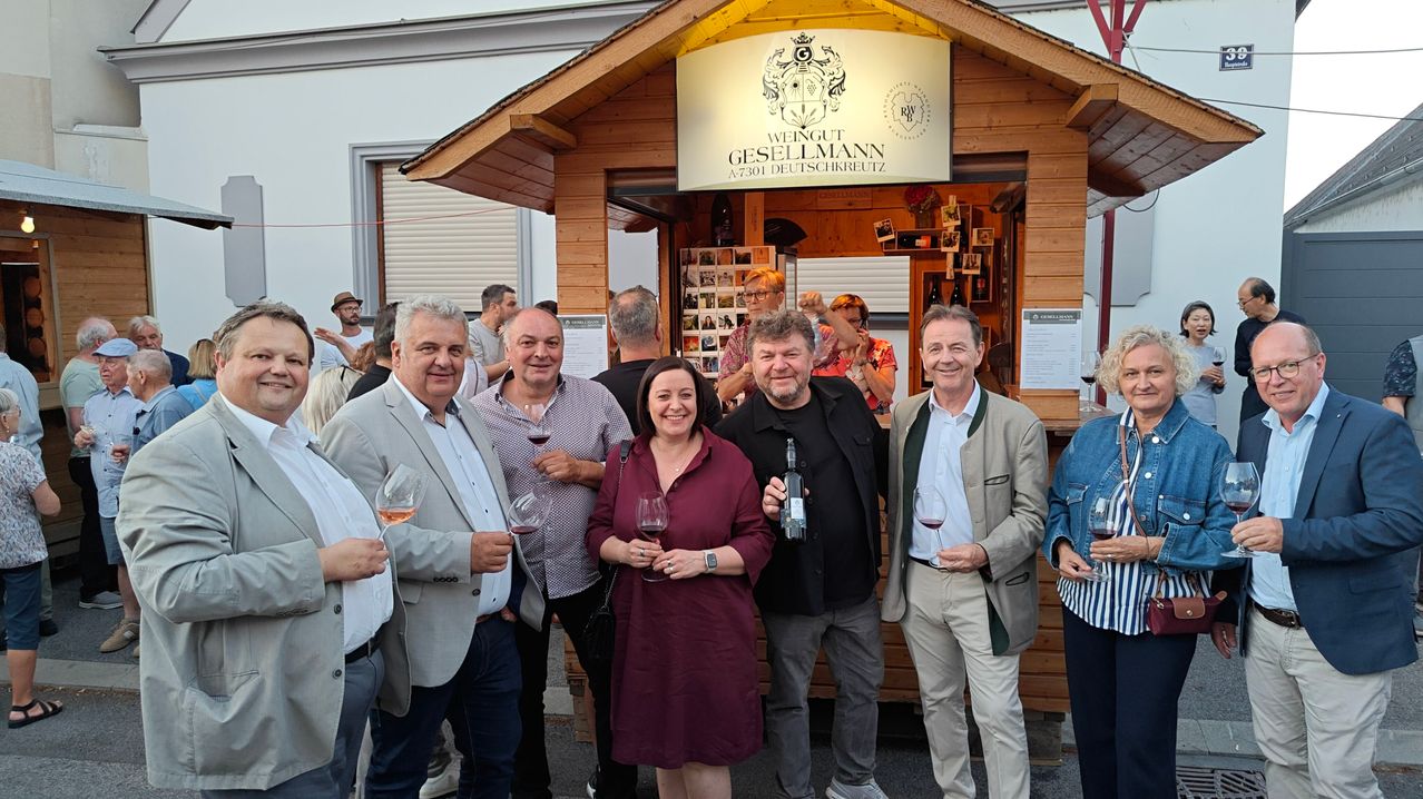A group of people are standing in front of a wooden booth with a sign that reads 'Weingut Gesellmann'. They are all smiling and holding wine glasses. The man in the middle is holding a bottle of wine. Behind them is a blurry background of people and a building.