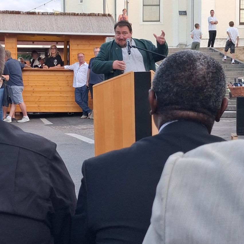 A man speaks at a podium while others watch from a wooden booth and steps.