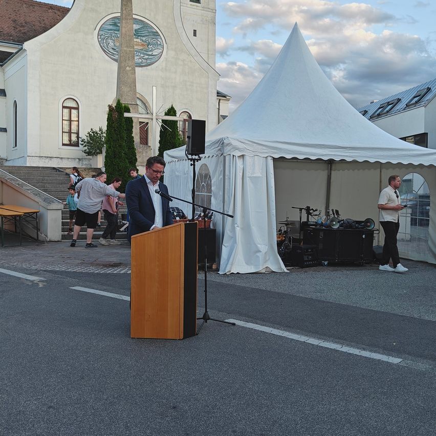 A man speaks at a podium in front of a church with a large white tent set up nearby.