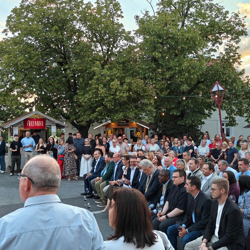 A large group of people are gathered in an outdoor area, possibly for an event. Some are seated on chairs, while others stand around. The area is surrounded by trees and a building with a red sign. The sky is blue with some clouds.