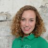 A smiling woman with curly hair wearing a green polo shirt with a logo on the left chest area. The background is a stone wall.