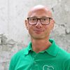 Ein kahlköpfiger Mann, der ein grünes Polohemd mit einem weißen Herzlogo trägt, lächelt und trägt eine Brille.