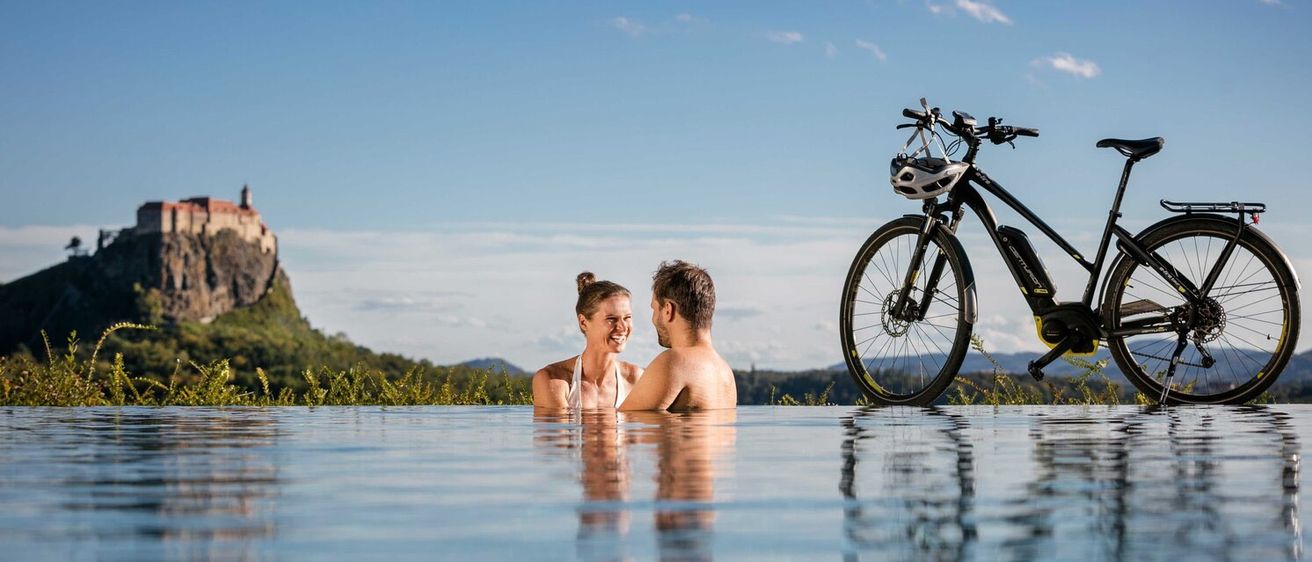 Ein Paar entspannt sich in einem Pool, mit ihrem Elektrofahrrad im Hintergrund, unter einem klaren Himmel mit entfernten Hügeln und einer Burg.