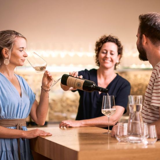 Eine Frau trinkt Wein, während eine Kellnerin ihr und einem Mann Wein einschenkt.