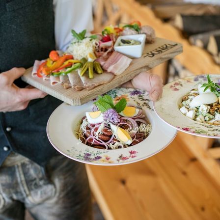 Ein Kellner hält ein Holzbrett mit verschiedenen Fleischsorten und einem Holzschneidebrett mit Käse. Zwei Teller mit buntem Gemüse und Eiern sind in seinen Händen.