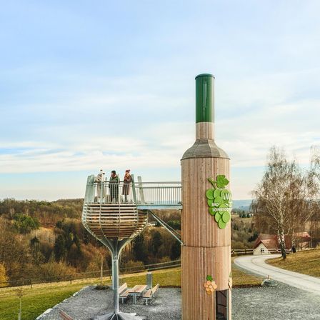 Ein Freiluftbereich mit einer großen flaschenförmigen Struktur, Menschen auf dem Balkon und Blick auf eine hügelige Landschaft.