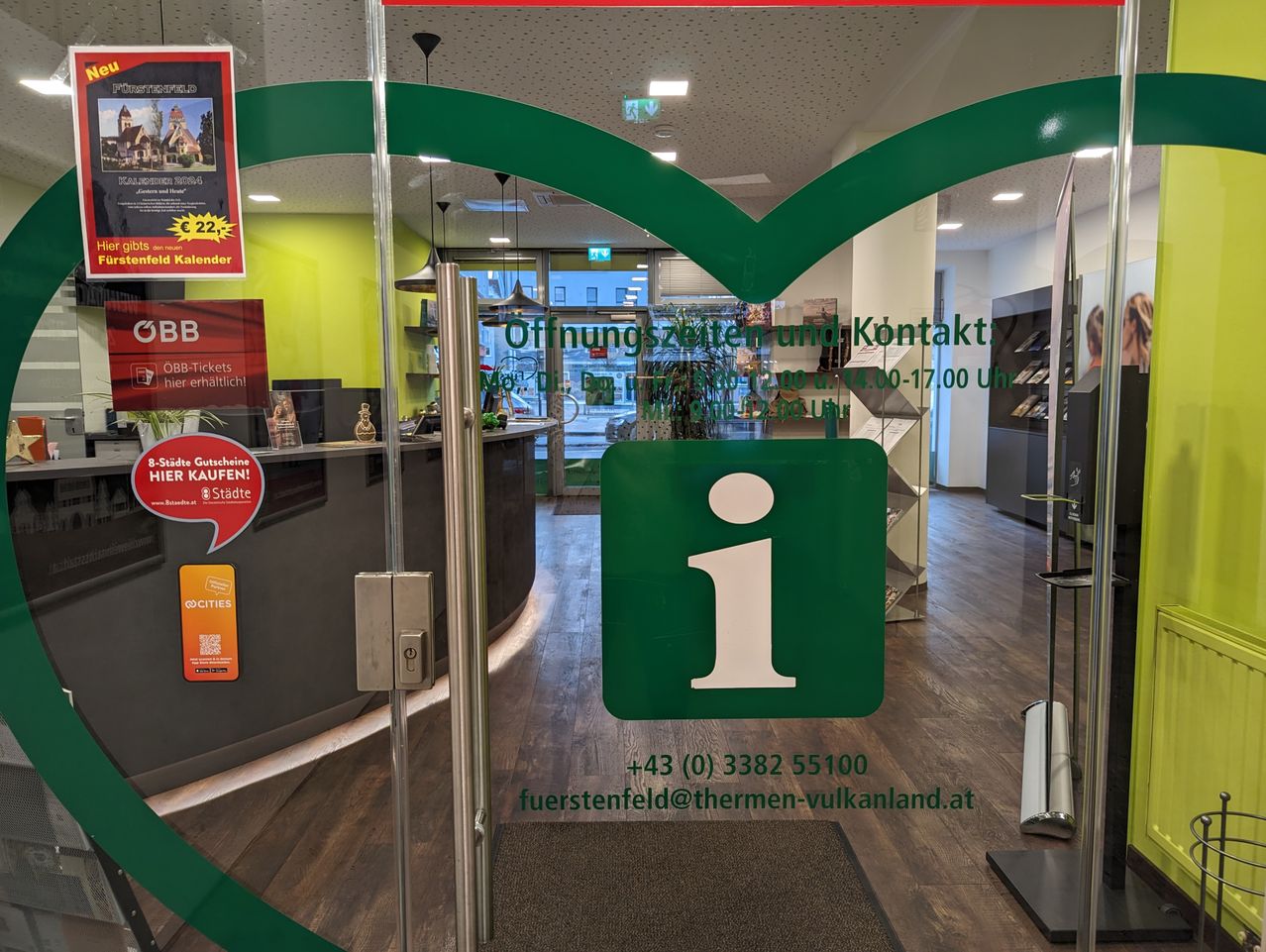 Das Bild zeigt eine Glastür mit grünen Akzenten, die Öffnungszeiten und Kontaktdaten anzeigt. Ein grünes 'i' ist prominent zu sehen. Im Inneren befindet sich ein Empfangsbereich mit einem Tresen und einigen Gegenständen. Der Boden ist aus Holz.