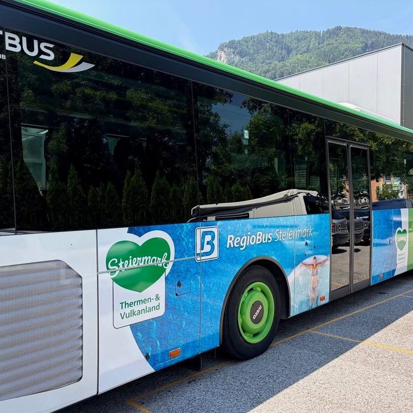 Ein RegioBus aus Steiermark mit einem grünen Herzlogo und dem Text 'Thermen- & Vulkanland' vor einem Gebäude mit Bergen im Hintergrund geparkt.