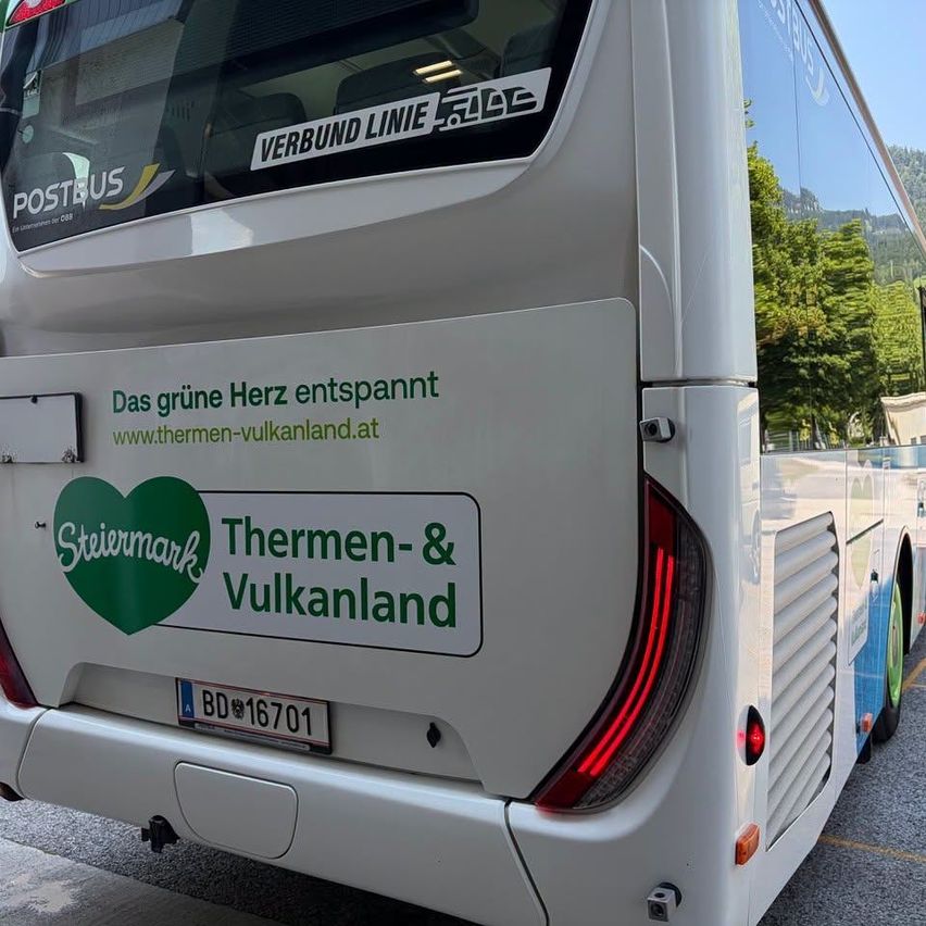 Die Rückseite eines weißen Busses mit einem grünen Herzlogo und der Aufschrift 'Thermen & Vulkanland'. Er hat ein Nummernschild 'BD 16701' und einen Aufkleber mit der Aufschrift 'Das grüne Herz entspannt www.thermen-vulkanland.at'. Der Bus ist am Straßenrand geparkt.