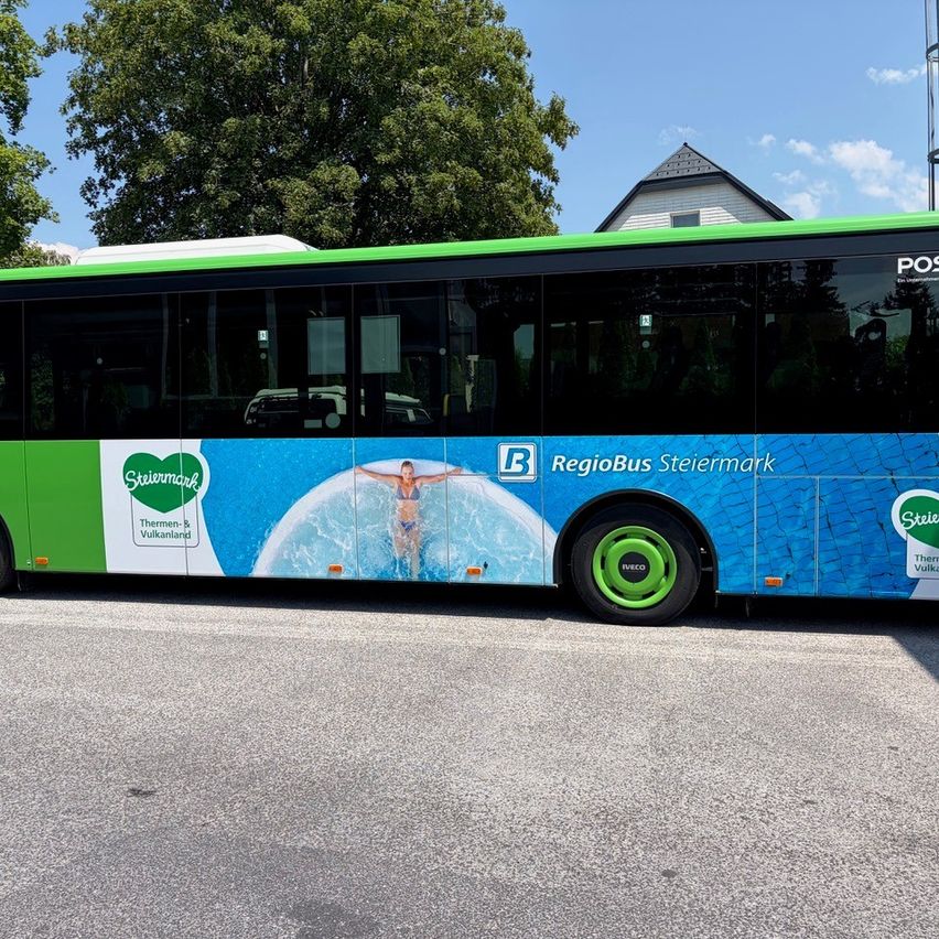 Ein grüner und blauer RegioBus von Steiermark steht am Straßenrand, mit einer Frau, die in einem Pool auf der Seite schwimmt.