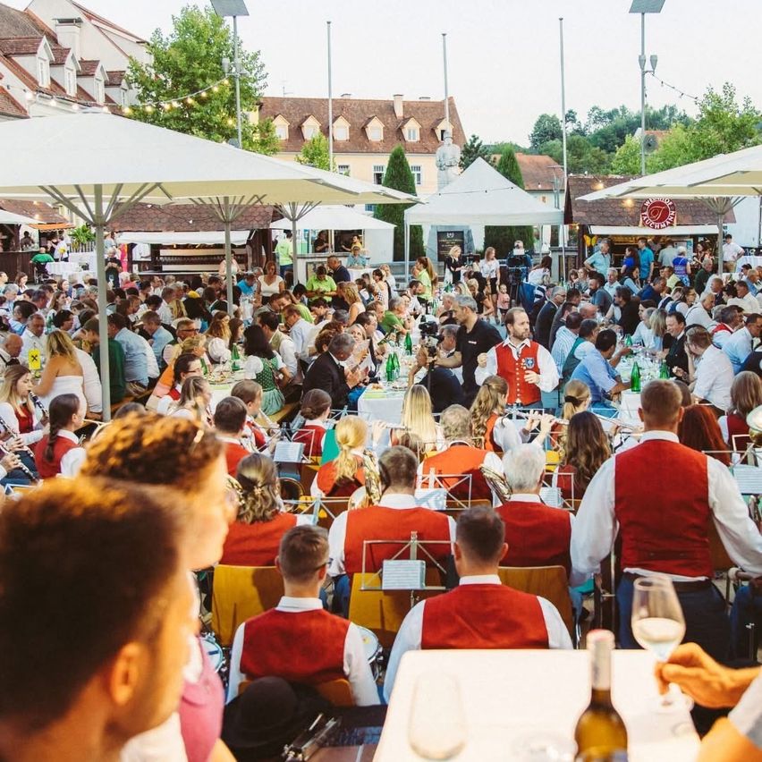 Eine große Menschenmenge versammelt sich im Freien, wahrscheinlich für ein Festival. Viele sitzen um Tische herum, einige stehen, und eine Band spielt im Vordergrund. Es gibt große Schirme, die Schatten spenden.