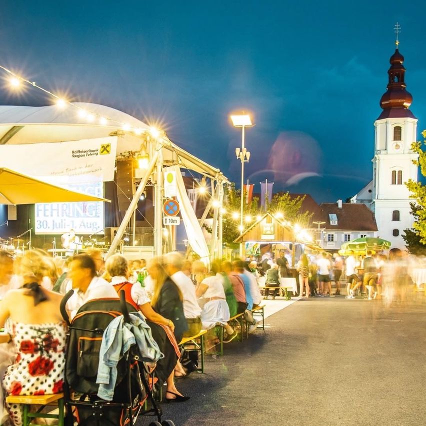 Ein geschäftiges Abendereignis mit Menschen, die unter einem Zelt sitzen und herumlaufen, mit einem Kirchturm und Straßenlaternen im Hintergrund.