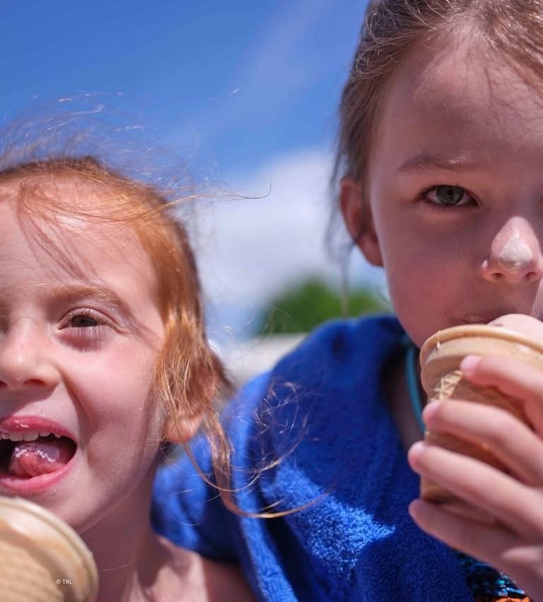 Zwei junge Mädchen, eines mit roten Haaren und das andere mit blonden, essen Eisbecher. Das Mädchen mit roten Haaren hat einen schelmischen Ausdruck.